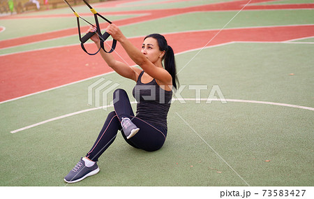 Girl athlete training using trx on sportground. Mixed race young adult woman do workout with suspension system. Healthy lifestyle. Stretching outdoors playground. 73583427