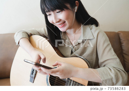 Happy asian woman in earphones listening to music and learning online guitar lesson with smartphone while sitting on couch at home. Hobbies and leisure activities concept. Happy asian woman in earphones listening to music and learning online guitar lesson with smartphone while sitting on couch at home. Hobbies and leisure activities concept. 73586283