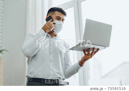 Handsome man businessman in medical mask using his laptop while talking on his cellphone Handsome man businessman in medical mask using his laptop while talking on his cellphone 73591510