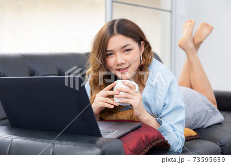 Beautiful young Asian woman enjoying her time at home, having her hot coffee while working on her computer on her sofa at home Beautiful young Asian woman enjoying her time at home, having her hot coffee while working on her computer on her sofa at home 73595639