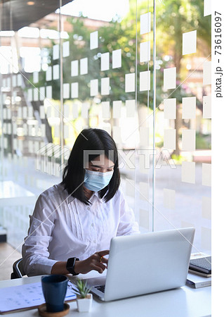 Young woman office worker in protective mask working or check information from website on her laptop at office. Young woman office worker in protective mask working or check information from website on her laptop at office. 73616097