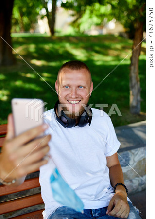 Man in medical mask sitting on the bench in the park take selfie by mobile phone. 73625750