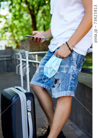 Man with protective mask in hand, standing in the park outdoor with a suitcase 73625813