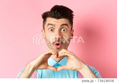 Valentines day concept. Close up of handsome caucasian man falling in love, looking amazed and showing heart gesture, pink background 73628056