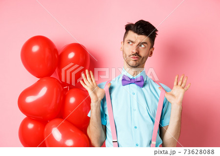 Valentines day concept. Pensive young man in bow-tie, raising hands up and looking left while thinking, making decision, standing on romantic pink background and heart balloons Valentines day concept. Pensive young man in bow-tie, raising hands up and looking left while thinking, making decision, standing on romantic pink background and heart balloons 73628088