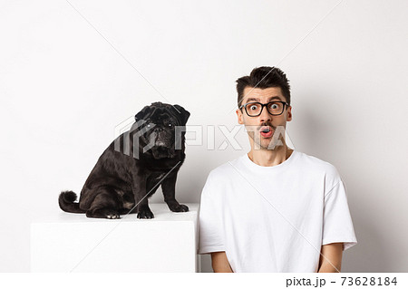 Portrait of dog owner and small cute pug staring at camera surprised and amazed, standing over white background Portrait of dog owner and small cute pug staring at camera surprised and amazed, standing over white background 73628184