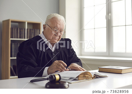 Serious wise mature judge in gown sitting at desk in courtroom and reading a book 73628956