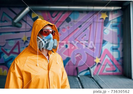 Graffiti painter in respirator mask standing near the wall with his paintings Graffiti painter in respirator mask standing near the wall with his paintings 73630562
