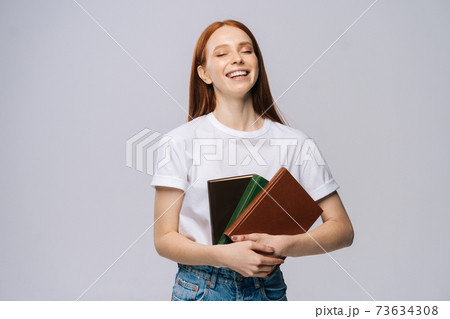 Laughing young woman college student with closed eyes holding books on isolated gray background. 73634308