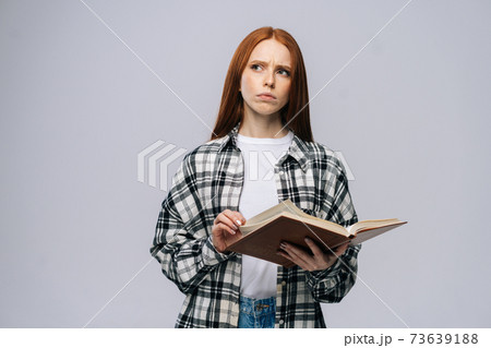 Thoughtful young woman college student turning pages of book on gray isolated background. 73639188