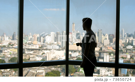 Dreaming european man stands by the window with great views and drinks hot coffee or tea, enjoying beautiful morning feeling happy Dreaming european man stands by the window with great views and drinks hot coffee or tea, enjoying beautiful morning feeling happy 73650724