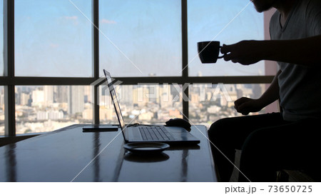 A concentrated young businessman is working on a laptop computer using his notebook sitting by the window with urban view and drinks coffee 73650725