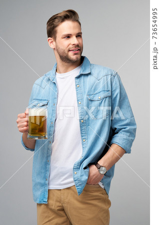 Young Man holding wearing jeans shirt holding glass of beer standing over Grey Background 73654995