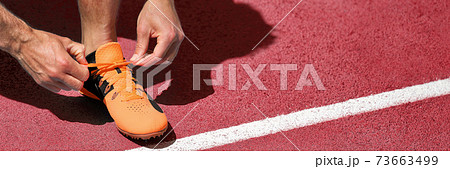 Running shoe at start line success challenge competition banner. Athlete fit man getting ready for run race tying running shoes. Panoramic on red tartan tracks. 73663499