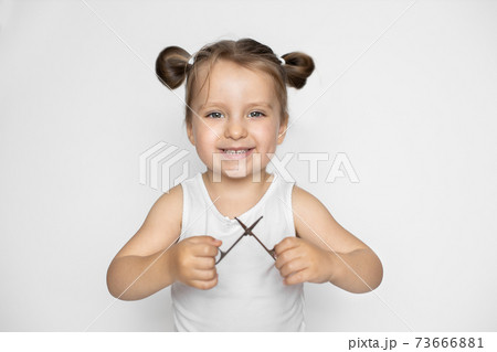 Baby manicure. Close up of cheerful little baby girl, wearing white top, holding in hands child-safe scissors for manicure. Close up on isolated white background. Copy space. 73666881