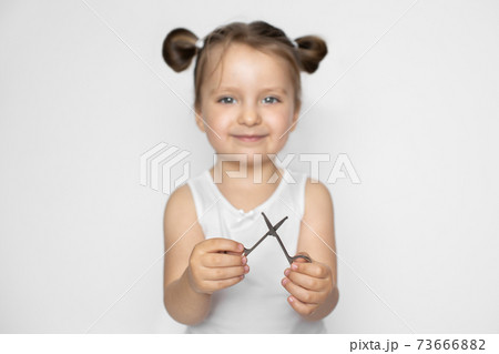 Happy smiling little girl with blonde hair holding manicure scissors, ready to trim her nails. Indoor studio portrait over white background, focus on scissors Happy smiling little girl with blonde hair holding manicure scissors, ready to trim her nails. Indoor studio portrait over white background, focus on scissors 73666882