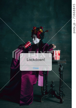 Young japanese woman as geisha isolated on dark green background. Drinking cola and eating popcorn behind limiting message lockdown on the screen 73668845