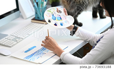 Close up view of Artist or designer woman with a palette in hands painting on paper. Close up view of Artist or designer woman with a palette in hands painting on paper. 73693188