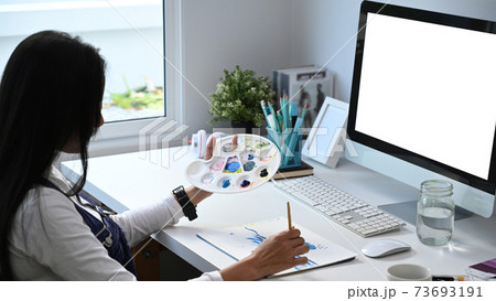 Young woman artist drawing painting with water color on paper in art studio while sitting in front of her computer. 73693191