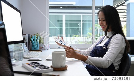 Side view of cheerful artist woman working on art projects at her workstation. Side view of cheerful artist woman working on art projects at her workstation. 73693192