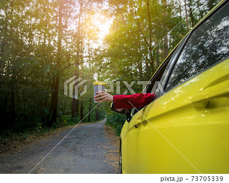 Young woman holds out a takeaway paper cup with hot drink through the driver's seat window. Focus on paper cup 73705339