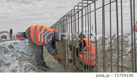 Engineers work with metal constructions while building a house. Builder fixes armature while working on construction site. Workers at joining metallic iron bars structure for reinforcement wall 73709190