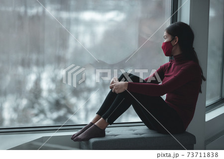 Winter depression because of coronavirus confinement. Sad Asian woman alone during city lockdown wearing face mask indoors at home covid. Anxiety, stress, mental health issues 73711838