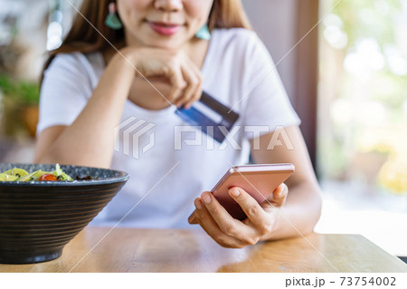 Young asian woman holding a credit card and using smart phone for make an online shopping payment at restaurant 73754002