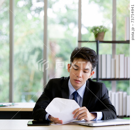 Young asian employee peeking inside the envelope. Morning atmosphere in the office on a table with tablet and laptop computer. 73754941