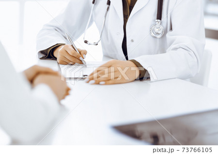 Unknown woman-doctor and female patient sitting and talking at medical examination in clinic, close-up. Therapist wearing green blouse is filling up medication history record. Medicine concept 73766105