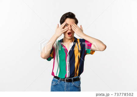 Image of handsome young gay man open eyes to see surprise, peeking through fingers with amazed emotion, standing over white background 73778502