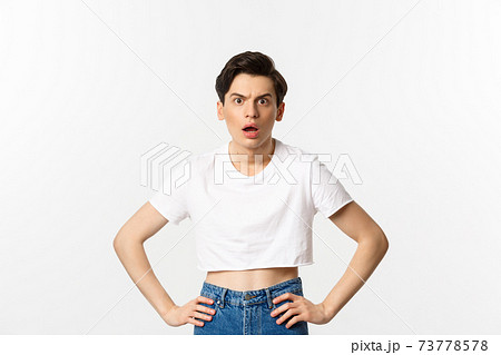 Shocked and confused gay man staring at camera startled, wearing crop top, holding hands on waist, standing over white background 73778578