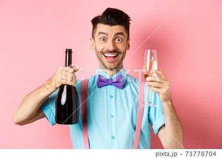 Holidays and celebration concept. Happy young man having fun, showing bottle of champagne and glass, making toast on event, smiling and looking at camera, pink background 73778704