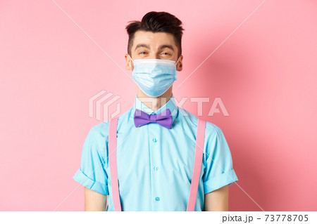 Covid-19, pandemic and health concept. Portrait of smiling man in medical mask feeling happy, standing in festive bow-tie and suspenders, pink background 73778705