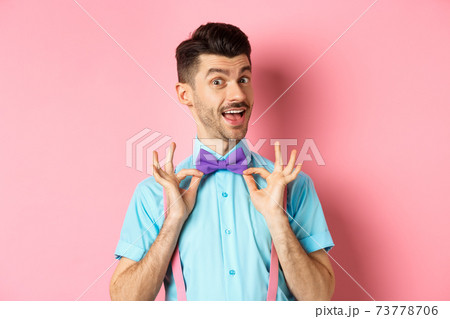 Classy handsome man fixing bow-tie on neck and smiling, getting ready for date or party, standing on pink background 73778706