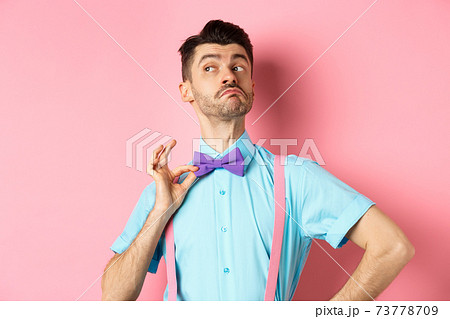 Image of funny man raising chin up and adjusting bow-tie, standing confident and sassy on pink background, looking left 73778709