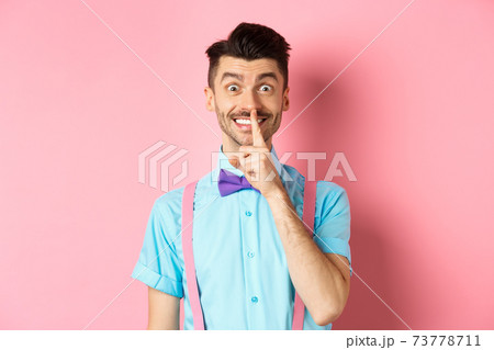 Cheerful young man making surprise, shushing at camera with happy smile, asking to keep voice down, be quiet, standing on pink background 73778711