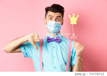 Covid, pandemic and quarantine concept. Disappointed guy in medical mask showing party crown and thumbs down, complaining while standing on pink background 73778712