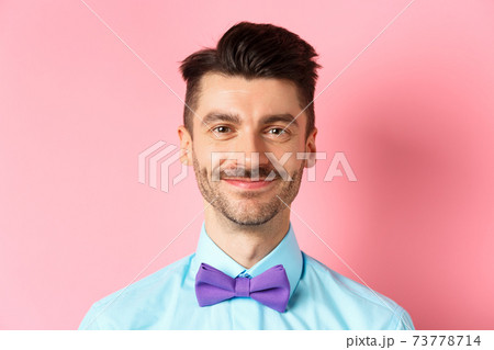 Close-up of smiling hansome man with moustache, wearing bow-tie and shirt, standing cheerful on pink background 73778714