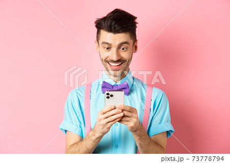 Online shopping. Happy guy looking at smartphone screen, reading message and smiling, standing on pink background 73778794