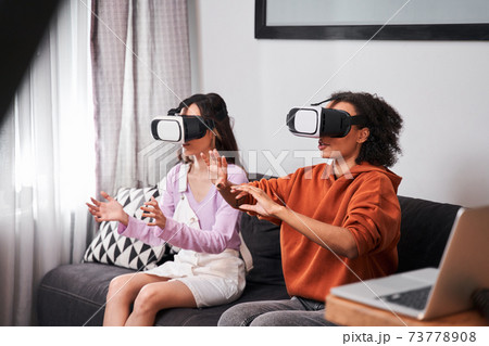 Besties sitting on cozy sofa and using virtual reality goggles Besties sitting on cozy sofa and using virtual reality goggles 73778908