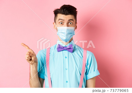 Covid-19, pandemic and health concept. Image of surprised guy in medical mask asking about promo deal, pointing finger left and look curious, standing over pink background 73779065