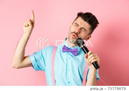 Party and festive events concept. Funny guy singing in microphone, raising finger up as reaching high note in song, standing on pink background Party and festive events concept. Funny guy singing in microphone, raising finger up as reaching high note in song, standing on pink background 73779079