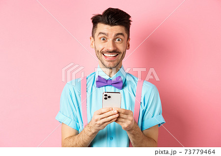 Online shopping. Happy guy in bow-tie, smiling at camera and using smartphone, standing on pink background 73779464