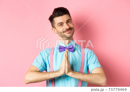 Smiling young guy in bow-tie say thank you, looking with gratitude and holding hands in namaste gesture, standing over pink background 73779469
