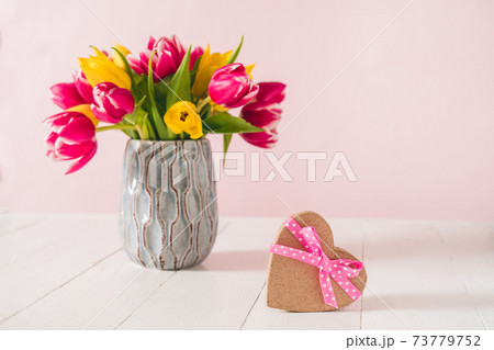 Craft heart shape gift box with satin ribbon bow and spring bouquet of pink and yellow tulip flowers in a vase on the white wooden table and pink background. Greeting card mockup. Copy space. Craft heart shape gift box with satin ribbon bow and spring bouquet of pink and yellow tulip flowers in a vase on the white wooden table and pink background. Greeting card mockup. Copy space. 73779752
