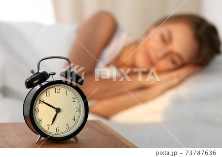 Beautiful young woman sleeping and smiling while lying in bed comfortably and blissfully on the background of alarm clock is going to ring. Sunbeam dawn on her face. Beautiful young woman sleeping and smiling while lying in bed comfortably and blissfully on the background of alarm clock is going to ring. Sunbeam dawn on her face. 73787636