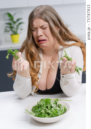 Fat girl not happy holding fresh salad casting. Curvy body young woman with long blond hair sitting on modern kitchen. Dieting and nutrition concept Fat girl not happy holding fresh salad casting. Curvy body young woman with long blond hair sitting on modern kitchen. Dieting and nutrition concept 73787815