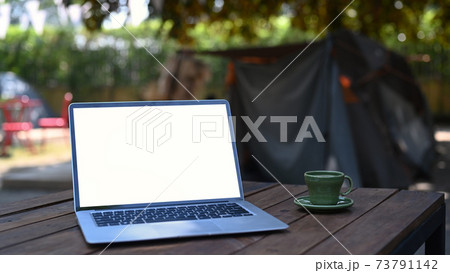 Laptop computer with blank screen and coffee cup on wooden table near camping tent. 73791142