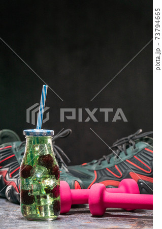 Fruit infuser water bottle with straw and fruits in water near trainers and dumbbells 73794665